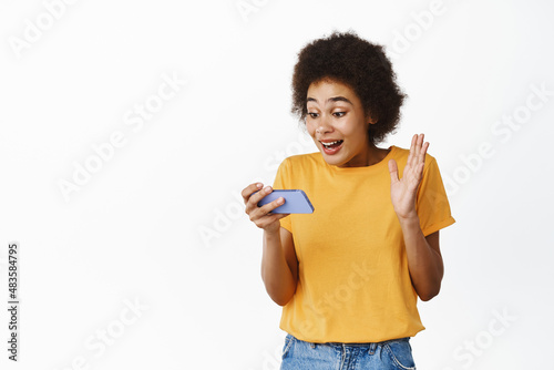 Portrait of black girl looking at her mobile phone with surprised, enthusiastic face expression, receive promo offer in app, standing over white background
