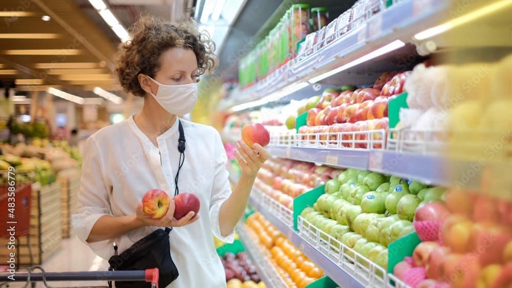 Female customer in protective mask choosing fresh red apples in groceries, fruits shopping 