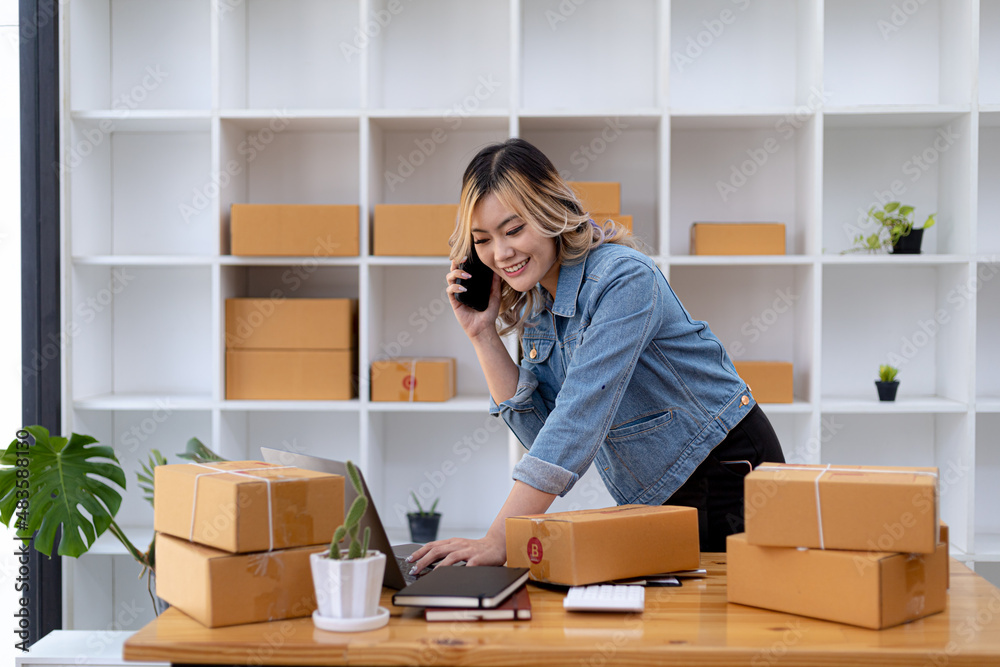 Asian woman is talking on the phone with a customer to confirm an order, she owns an online store, she packs and ships through a private transport company. Online selling and online shopping concepts.