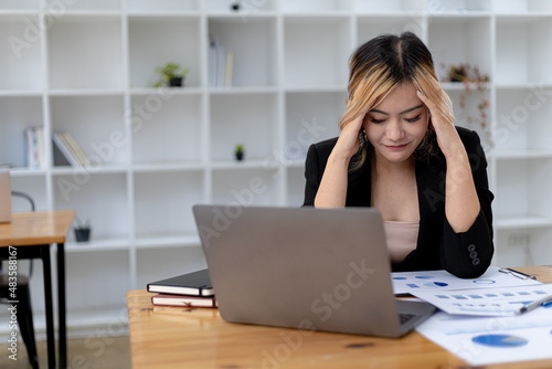 A businesswoman has a headache after working in front of her laptop for a long time and looking at a lot of paperwork causing stress, she is resting and working hard. Office syndrome concept. © kamiphotos