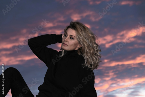 portrait of blonde woman dressed in black at sunset sitting on a wall touching her hair