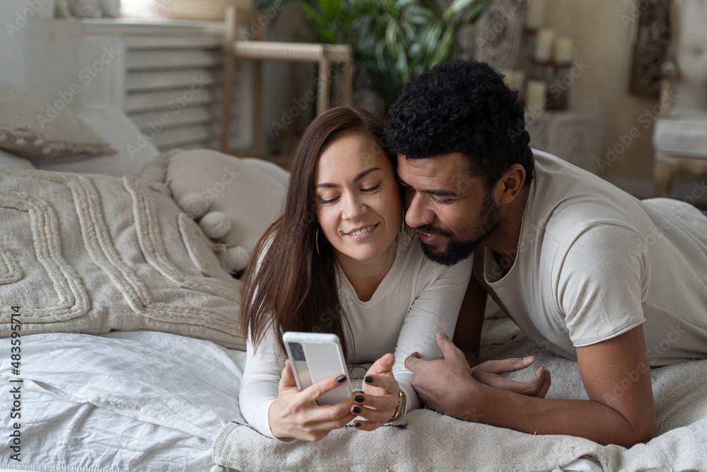 Smiling attractive couple using smartphone lying on bed together, happy guy and girlfriend checking social media news in morning, young woman showing man new mobile app Smiling attractive couple using smartphone lying on bed together, happy guy and girlfriend checking social media news in morning, young woman showing man new mobile app