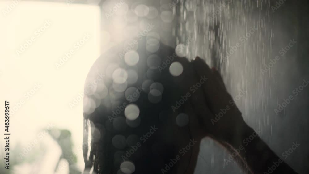 Black haired woman taking a shower and washing her face and hair while the condensation drops in front of the shower cubicle find their way down. Close up shot