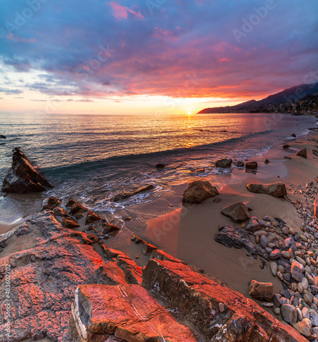 Sanremo, Riviera dei Fiori, Liguria, Italy. Scenis rocks and pebbles on beach illuminated beautiful by sunset light. Dramatic colourful sky waving sea.