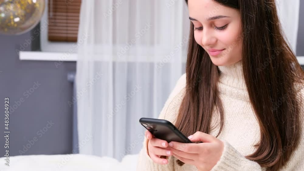 Happy smiling girl at home in white warm sweater using smart phone to chatting with her boyfriend, waiting to meet him. Happy smiling girl at home in white warm sweater using smart phone to chatting with her boyfriend, waiting to meet him.