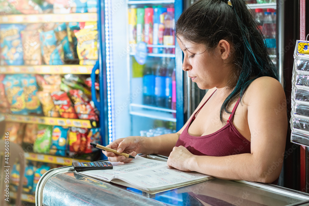 latina woman concentrating while calculating the number of sales in a notebook of her grocery store. girl leaning on a glass display case thinking about sales. concept of economy and business