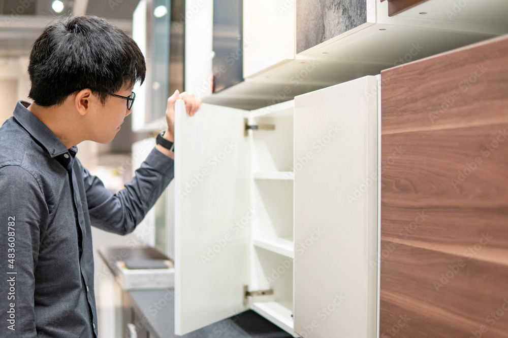 Asian man opening wooden cabinet door in furniture store choosing material to buy for house decoration. Home improvement concept Asian man opening wooden cabinet door in furniture store choosing material to buy for house decoration. Home improvement concept
