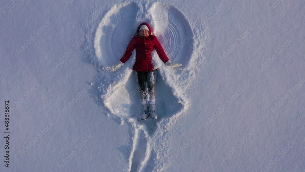 Aerial view from drone of girl in red winter coat making snow angel. Girl waves arms and legs lying in snow. High quality 4k footage