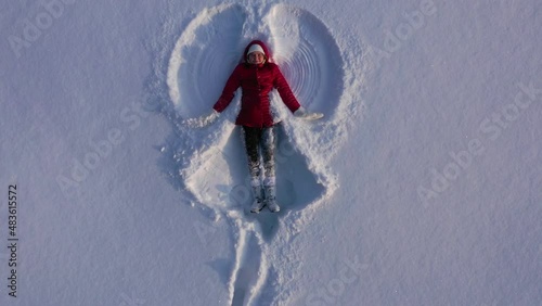 Aerial view from drone of girl in red winter coat making snow angel. Girl waves arms and legs lying in snow. High quality 4k footage photo
