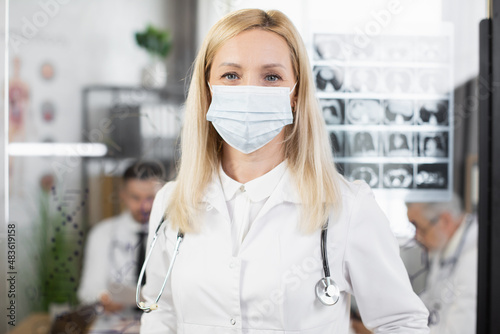 Caucasian female doctor in face mask and lab coat posing at conference room with transparent flip chart on background. Portrait of qualified medical worker.