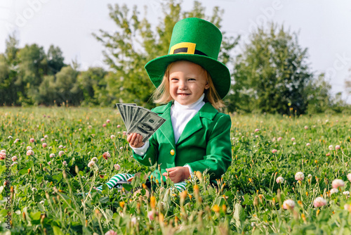 St. Patrick's Day holiday. A girl dressed in a Leprechaun costume sits in a clover meadow and holds $100 bills in her hands. Happy joyful toddler baby