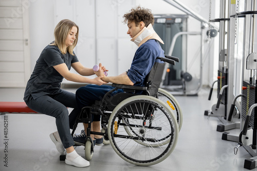 Photos Rehabilitation specialist helps a guy to do exercises for recovery from injury, who is sitting in a wheelchair with a corset around his neck