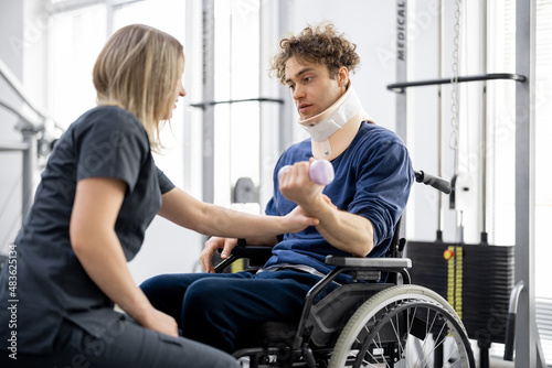 Rehabilitation specialist helps a guy to do exercises for recovery from injury, who is sitting in a wheelchair with a corset around his neck. Concept of physical therapy for people with disabilities