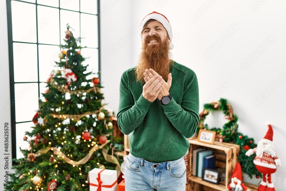 Redhead man with long beard wearing christmas hat by christmas tree suffering pain on hands and fingers, arthritis inflammation Redhead man with long beard wearing christmas hat by christmas tree suffering pain on hands and fingers, arthritis inflammation