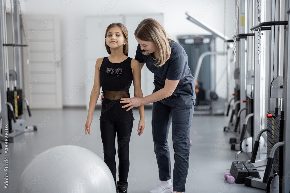 Rehabilitation specialist helping little girl to do exercises at gym. Concept of physical therapy for back health and correct posture for kids - obrazy, fototapety, plakaty 