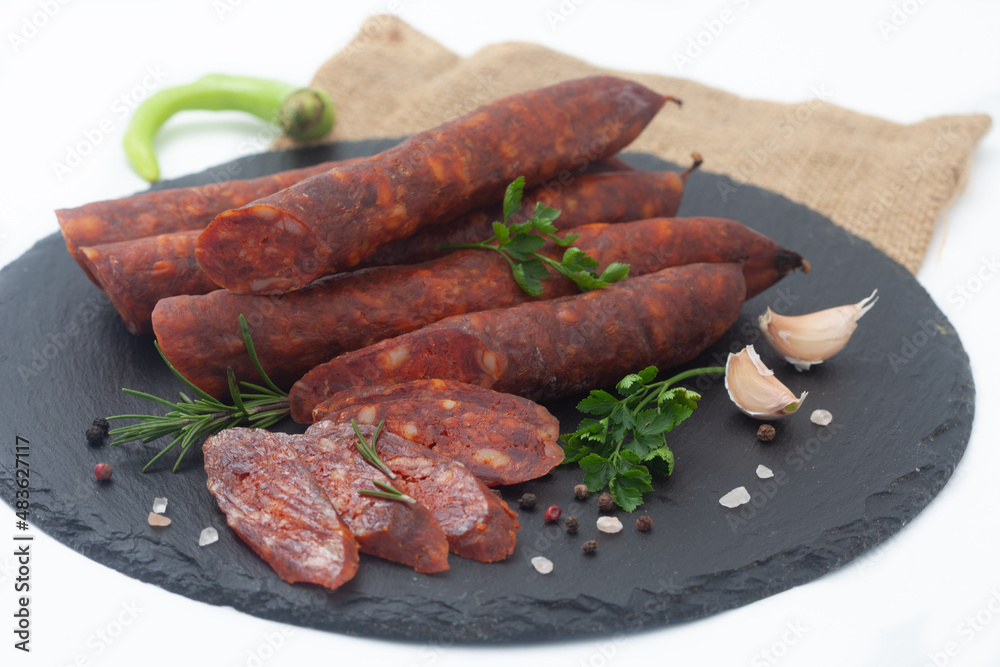 Dry sausages made of pork meat on a black board with fresh herbs and spices.  Smoked, homemade sausage on white background.