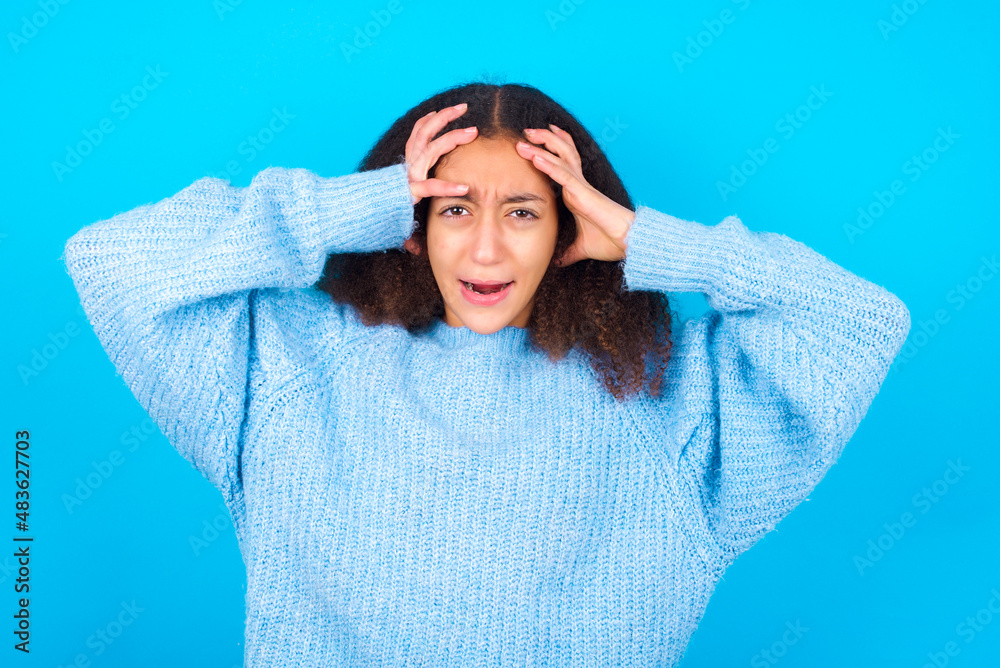 Shocked panic African teenager girl wearing blue sweater over blue background holding hands on head and screaming in despair and frustration. Shocked panic African teenager girl wearing blue sweater over blue background holding hands on head and screaming in despair and frustration.