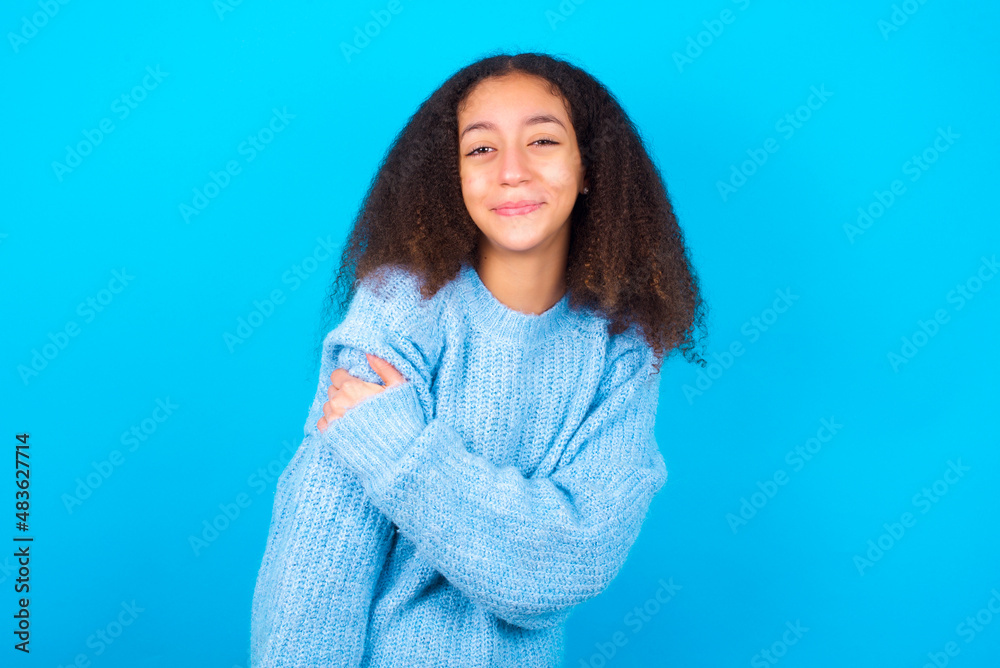 Fototapeta premium People, lifestyle, youth and happiness concept. Shy pretty African teenager girl wearing blue sweater over blue background, feeling happy hugging herself.