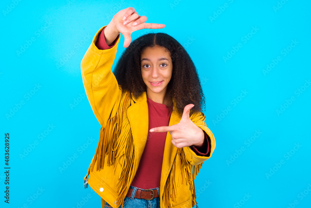Positive African teenager girl wearing yellow jacket over blue background with cheerful expression, has good mood, gestures finger frame actively at camera. Positive African teenager girl wearing yellow jacket over blue background with cheerful expression, has good mood, gestures finger frame actively at camera.