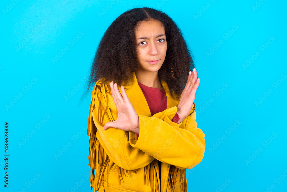 African teenager girl wearing yellow jacket over blue background Has rejection angry expression on face and crossing hands doing refusal negative sign. African teenager girl wearing yellow jacket over blue background Has rejection angry expression on face and crossing hands doing refusal negative sign.
