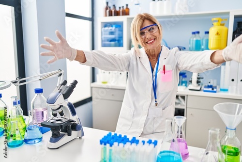 Middle age blonde woman working at scientist laboratory looking at the camera smiling with open arms for hug. cheerful expression embracing happiness.