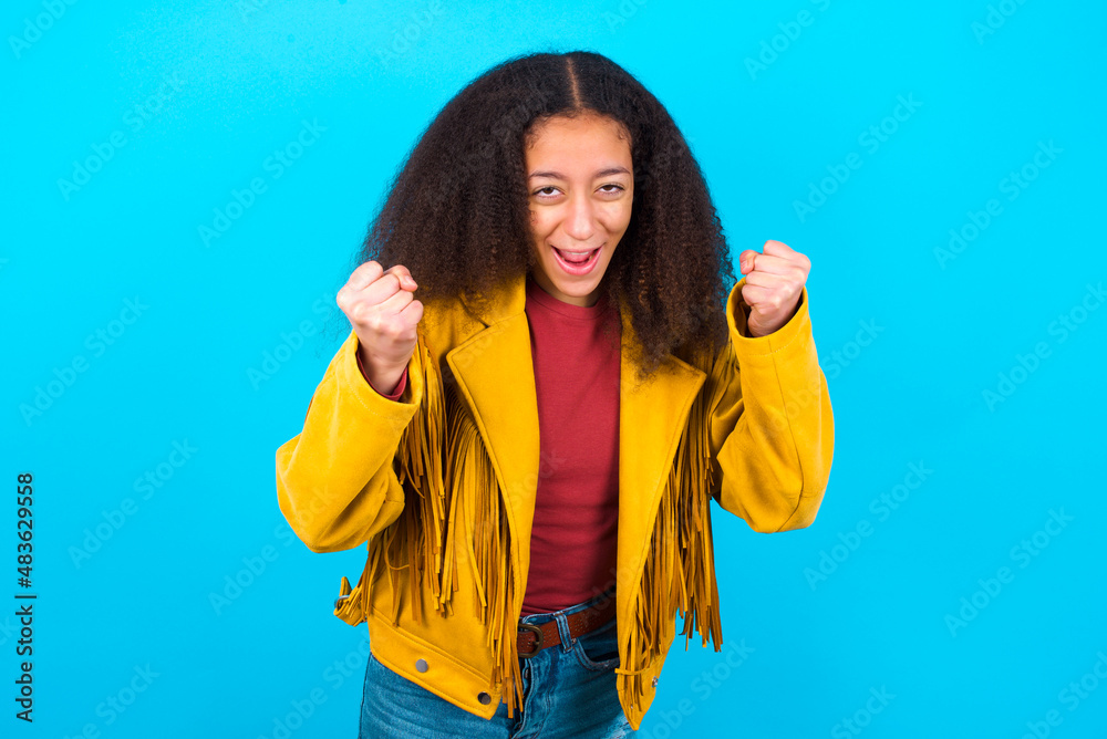 Portrait of African teenager girl wearing yellow jacket over blue background looks with excitement at camera, keeps hands raised over head, notices something unexpected reacts on sudden news.