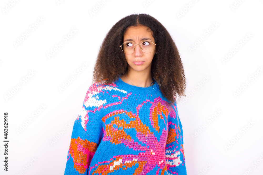 Dissatisfied African teenager girl wearing colorful sweater over white background purses lips and has unhappy expression looks away stands offended. Depressed frustrated model. Dissatisfied African teenager girl wearing colorful sweater over white background purses lips and has unhappy expression looks away stands offended. Depressed frustrated model.