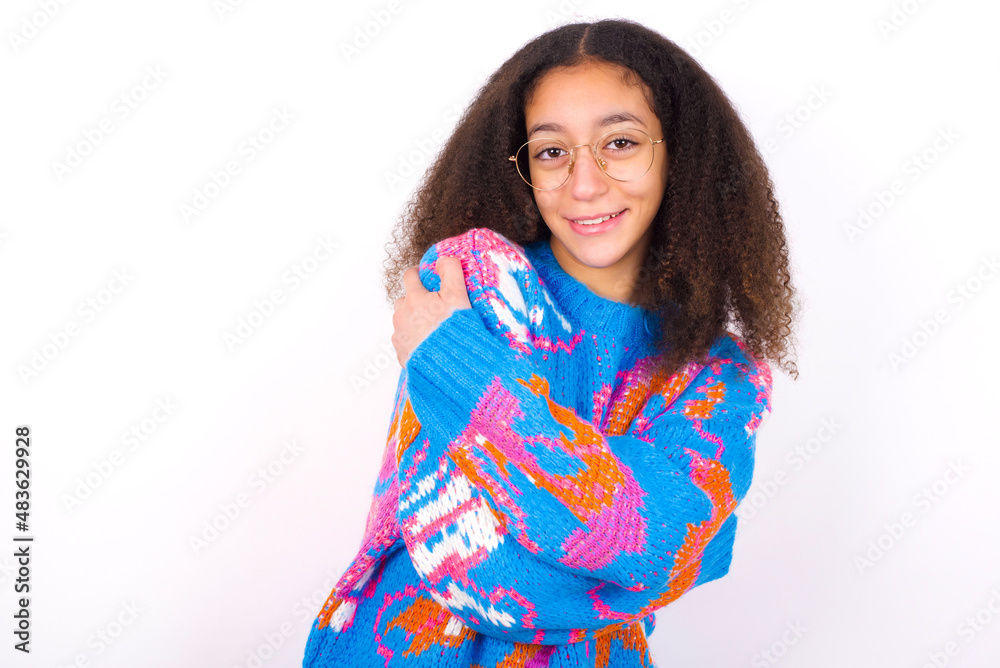 Charming pleased African teenager girl wearing colorful sweater over white background embraces own body, pleasantly feels comfortable poses. Tenderness and self esteem concept Charming pleased African teenager girl wearing colorful sweater over white background embraces own body, pleasantly feels comfortable poses. Tenderness and self esteem concept