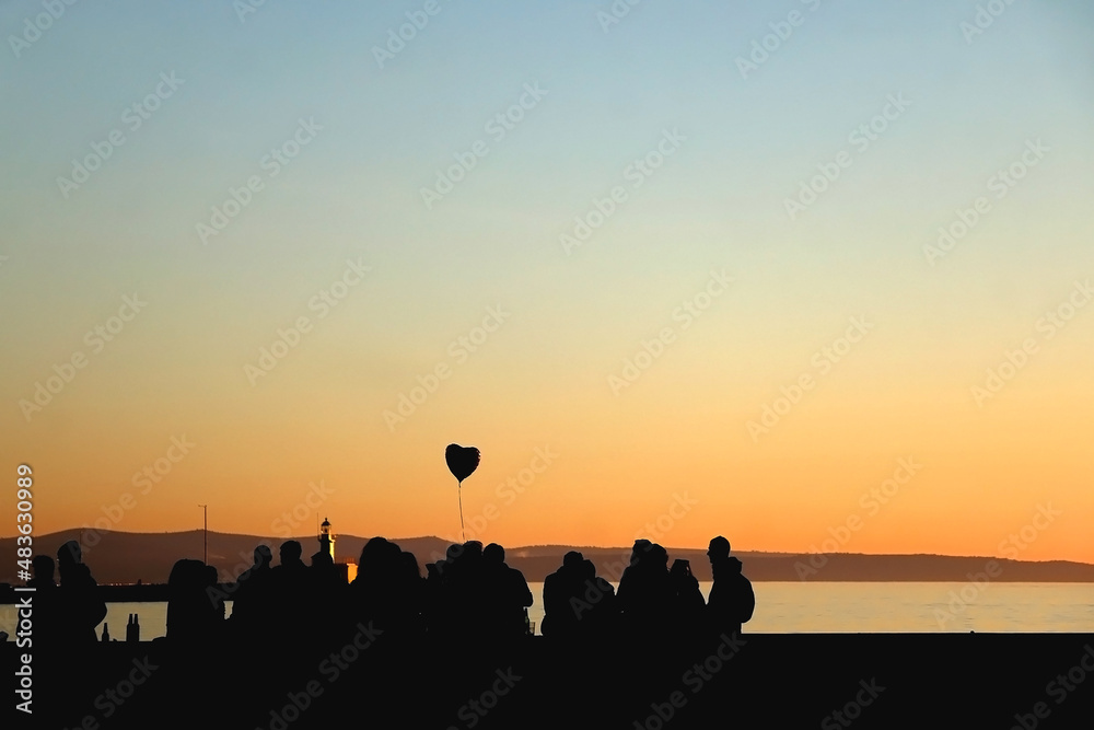 Silhouette of unrecognizable group of people and heart shaped balloon, enjoying sunset. Selective focus.