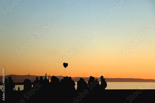 Silhouette of unrecognizable group of people and heart shaped balloon  enjoying sunset. Selective focus.