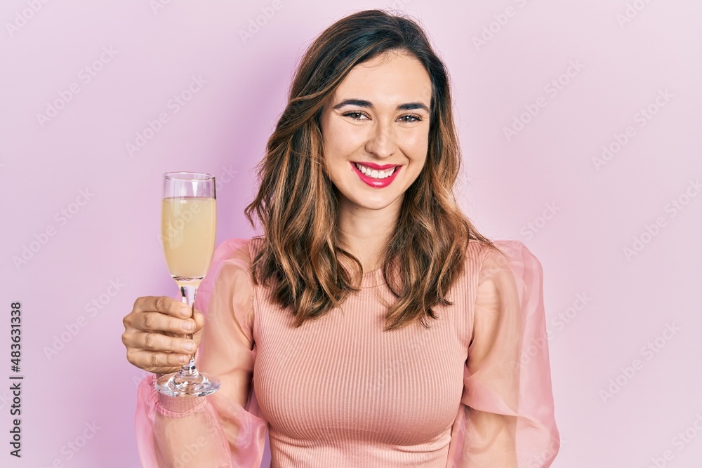 Young hispanic girl drinking a glass of sparkling champagne looking positive and happy standing and smiling with a confident smile showing teeth Young hispanic girl drinking a glass of sparkling champagne looking positive and happy standing and smiling with a confident smile showing teeth