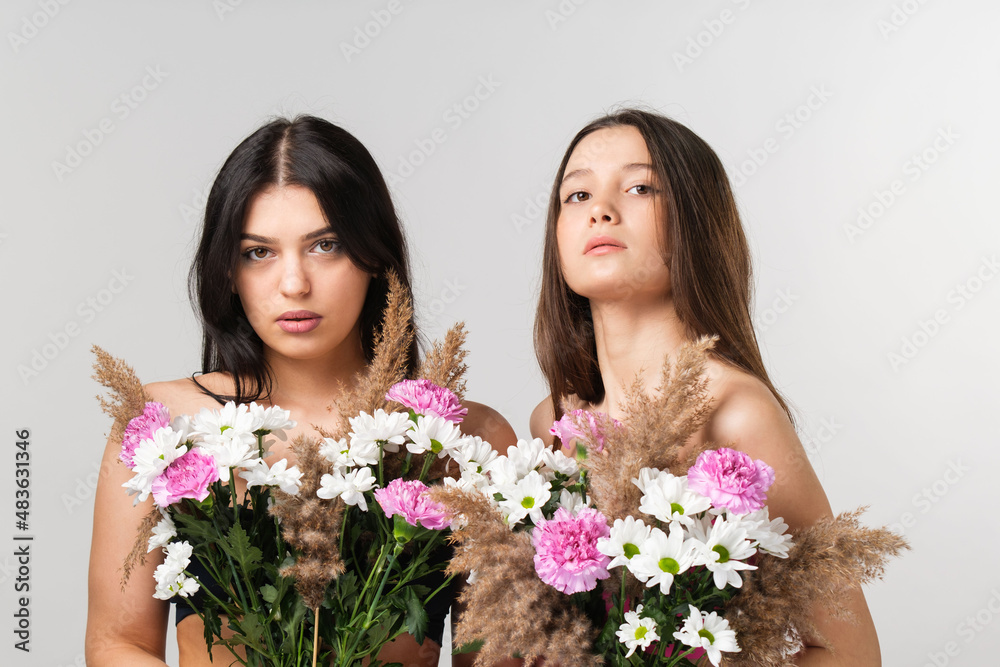 Fototapeta Zbliżenie na zdjęcie dwóch pięknych dziewczyn, blondynki i brunetki z nagimi ramionami zakrywającymi piersi kolorowym bukietem kwiatów.