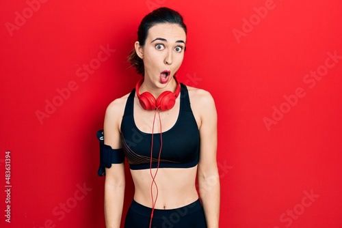 Young hispanic girl wearing gym clothes and using headphones afraid and shocked with surprise expression, fear and excited face.