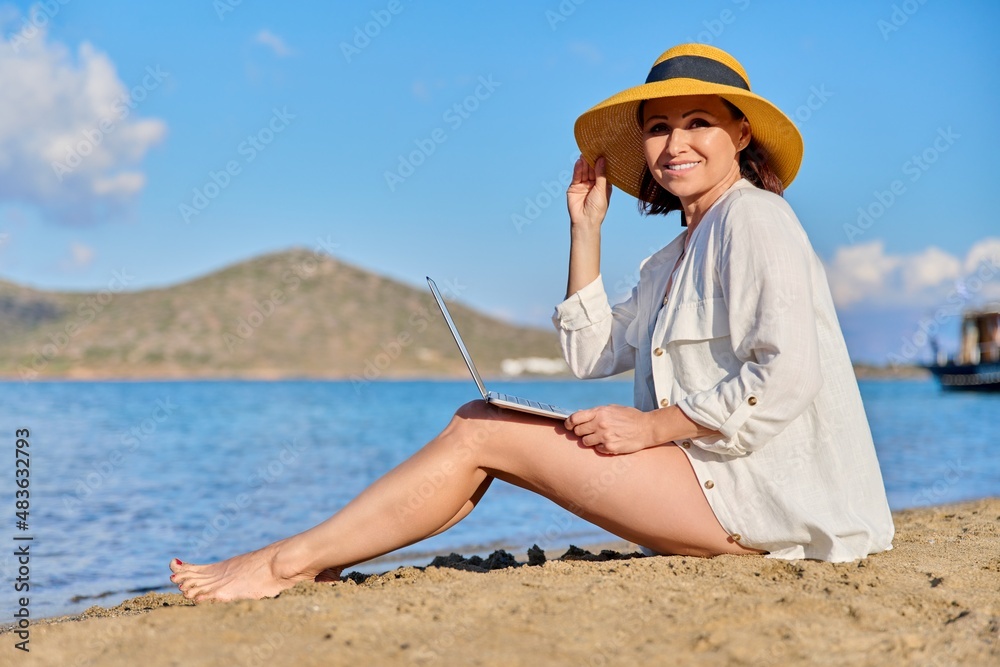 Mature woman in hat using laptop on sea beach