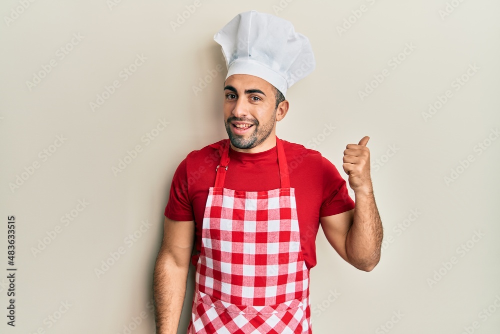 Young hispanic man wearing baker uniform smiling with happy face looking and pointing to the side with thumb up.