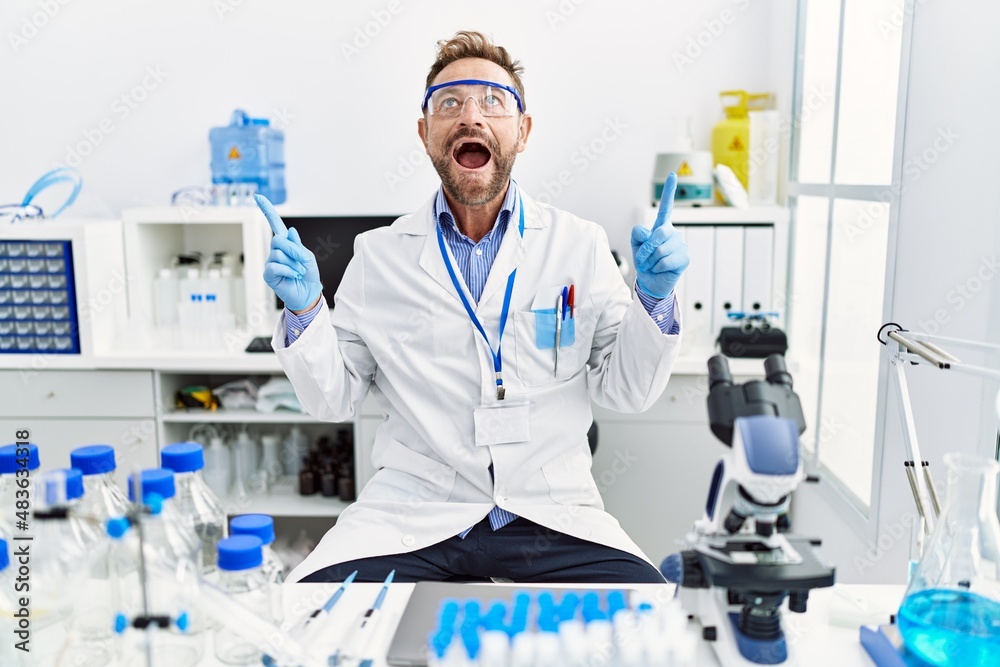 Middle age man working at scientist laboratory amazed and surprised looking up and pointing with fingers and raised arms. Middle age man working at scientist laboratory amazed and surprised looking up and pointing with fingers and raised arms.