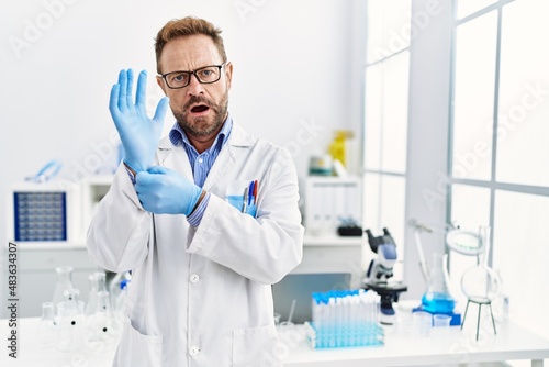 Middle age man working at scientist laboratory in shock face, looking skeptical and sarcastic, surprised with open mouth © Krakenimages.com