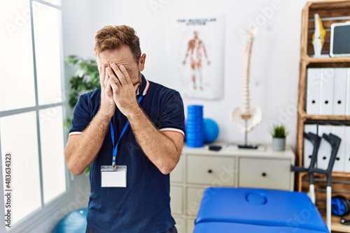 Middle age physiotherapist man working at pain recovery clinic with sad expression covering face with hands while crying. depression concept.