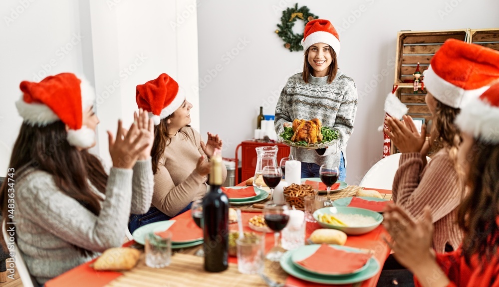 Fototapeta premium Group of hispanic women clapping and sitting on the table. Woman standing and holding roasted turkey celebrating Christmas at home.