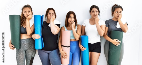 Group of women holding yoga mat standing over isolated background looking stressed and nervous with hands on mouth biting nails. anxiety problem.