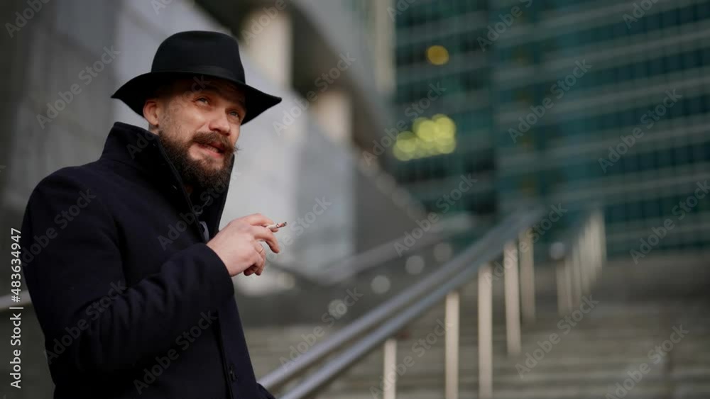 a mustachioed and bearded stylish man in a hat and a dark coat smokes a cigarette on the stairs against the background of a blurry modern building. bottom view a mustachioed and bearded stylish man in a hat and a dark coat smokes a cigarette on the stairs against the background of a blurry modern building. bottom view