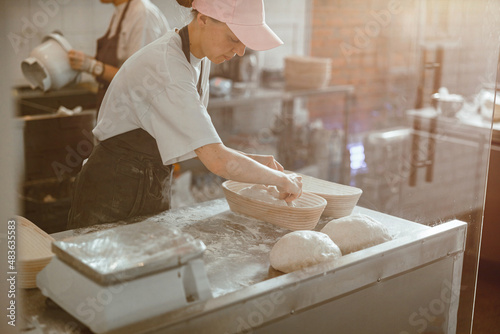 Employee in uniform decorates raw dough loaf in dish in bakery workshop