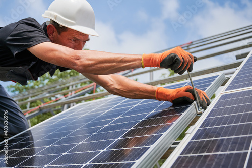 Close up of male worker mounting photovoltaic solar panel system outdoors. Man installer placing solar module on metal rails, wearing construction helmet and work gloves, tightening with hex key.