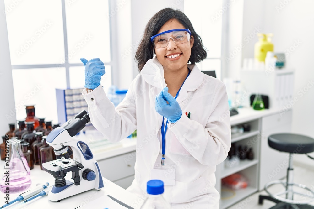 Young hispanic woman working at scientist laboratory with safety mask pointing thumb up to the side smiling happy with open mouth Young hispanic woman working at scientist laboratory with safety mask pointing thumb up to the side smiling happy with open mouth