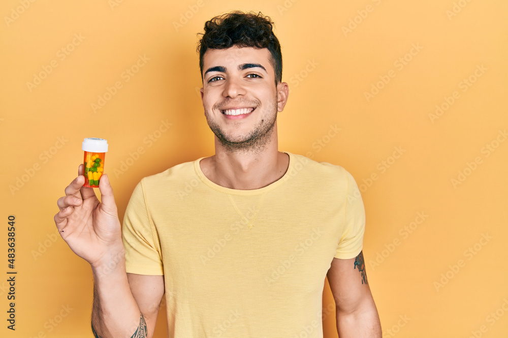 Young hispanic man holding pills looking positive and happy standing and smiling with a confident smile showing teeth Young hispanic man holding pills looking positive and happy standing and smiling with a confident smile showing teeth