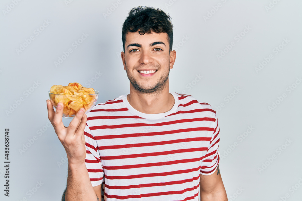 Young hispanic man holding potato chip looking positive and happy standing and smiling with a confident smile showing teeth