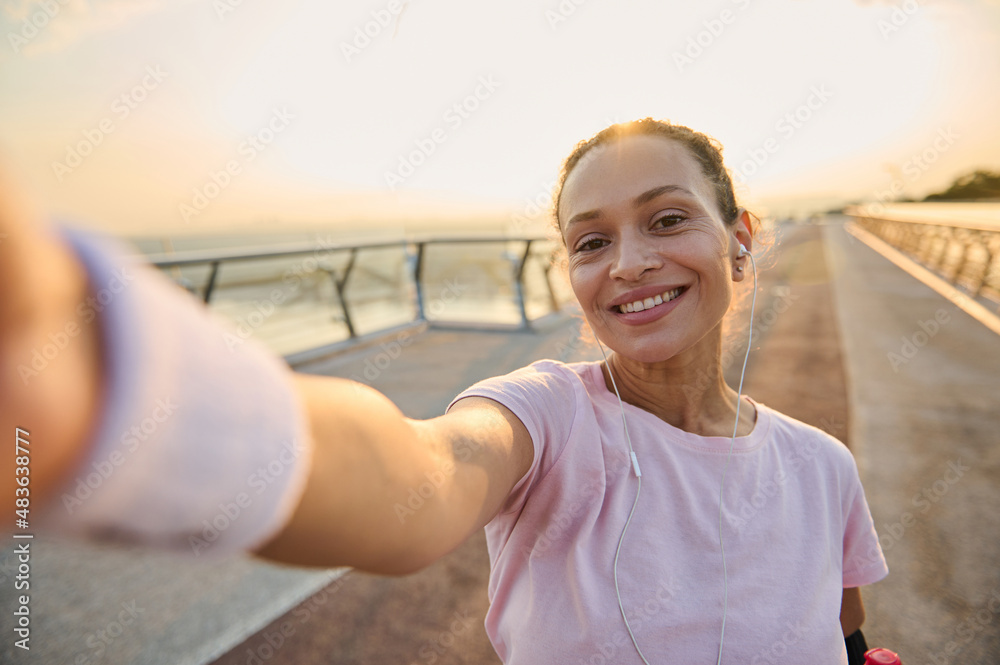 Sportswoman, female athlete in pink t-shirt and wristbands smiles holding a mobile phone in her outstretched hands while making self-portrait during morning jog and workout on the bridge at sunrise