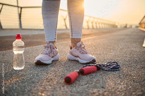 Croppped image of athlete legs wearing pink sneakers and standing on an asphalt treadmill next to a lying down jumping rope and water bottle against the background of a beautiful sunrise on the bridge