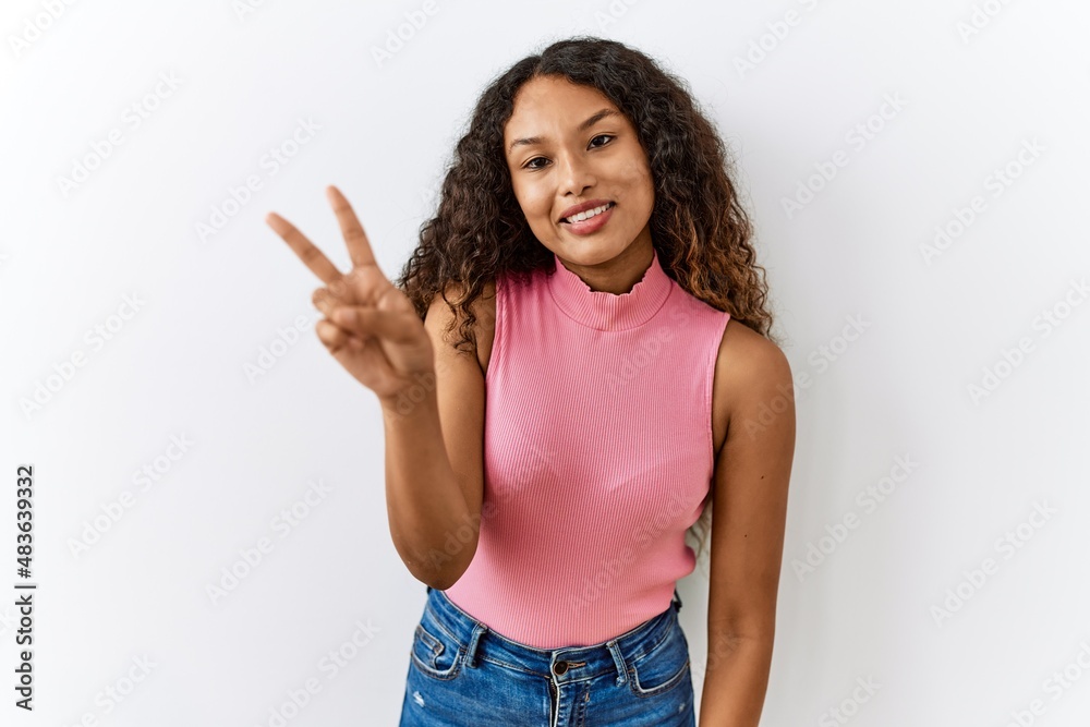 Beautiful hispanic woman standing over isolated background smiling looking to the camera showing fingers doing victory sign. number two. Beautiful hispanic woman standing over isolated background smiling looking to the camera showing fingers doing victory sign. number two.