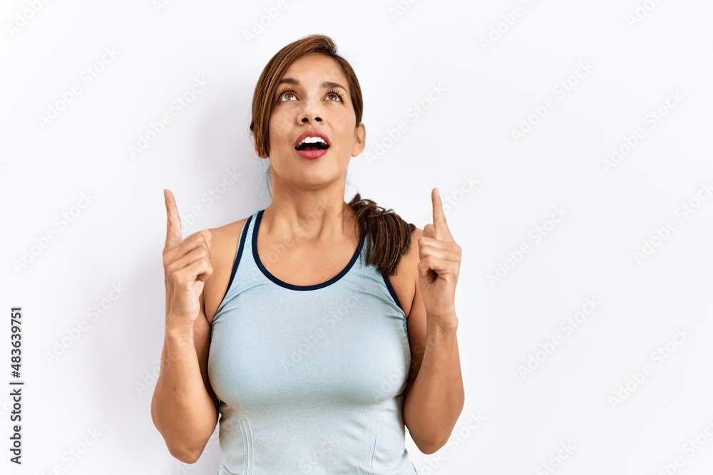 Young latin woman wearing sporty clothes over isolated background amazed and surprised looking up and pointing with fingers and raised arms. Young latin woman wearing sporty clothes over isolated background amazed and surprised looking up and pointing with fingers and raised arms.